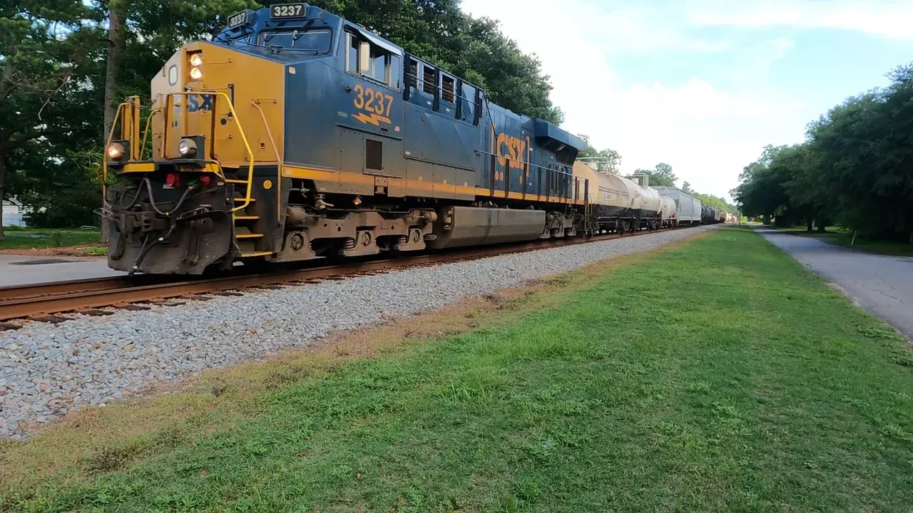 CSX locomotive 3237 pulling tank cars along track beside grassy path and trees